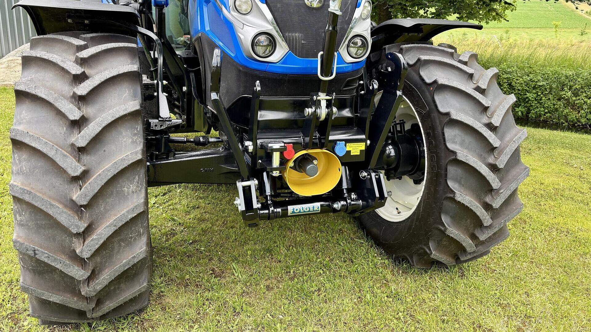 Nahaufnahme der Vorderseite eines blauen Traktors mit großen, robusten Reifen, der auf einer Wiese geparkt ist und dessen Maschinen und Anbauteile zeigt. Im Hintergrund sind Bäume und Sträucher zu sehen.