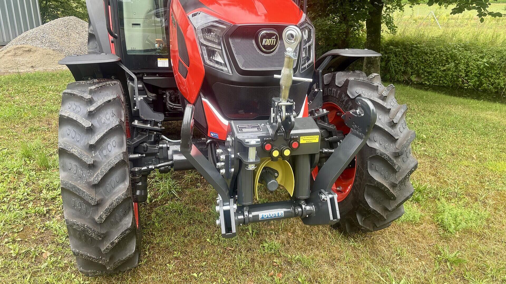 Ein roter Kioti-Traktor ist auf einer Wiese geparkt, mit Blick nach vorne. Der Traktor hat große, robuste Reifen, einen sichtbaren Frontkraftheber und ist von Grünflächen und Bäumen im Hintergrund umgeben.
