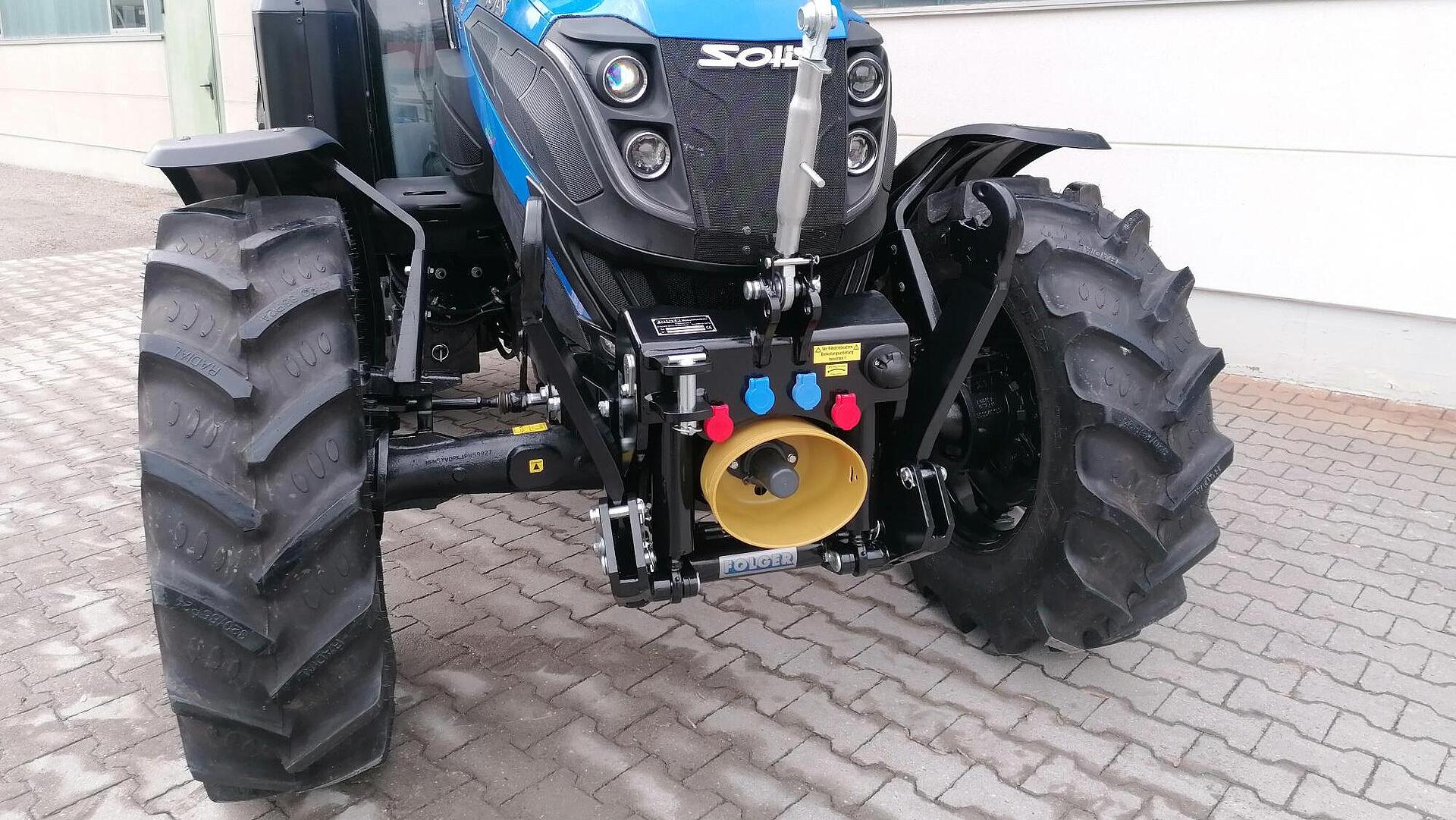 Ein blauer Solis-Traktor ist auf einer gepflasterten Fläche in der Nähe eines Gebäudes mit großen Fenstern geparkt, wobei die Vorderreifen, die hydraulischen Anschlüsse und der Frontanbau zu sehen sind.