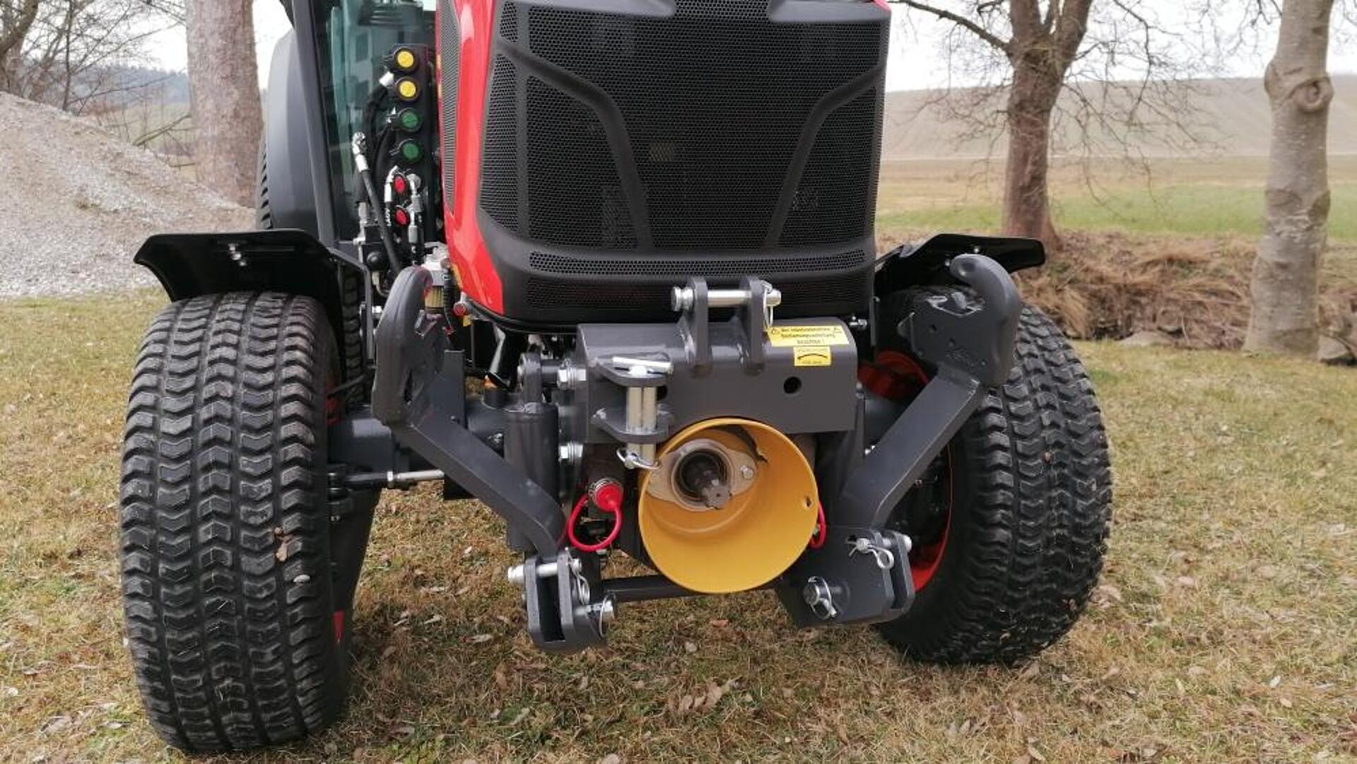 Vorderansicht eines roten Traktors, der auf einer Wiese geparkt ist, mit großen Reifen, Frontkupplung und gelber Zapfwelle; im Hintergrund sind Bäume und ein Feld zu sehen.