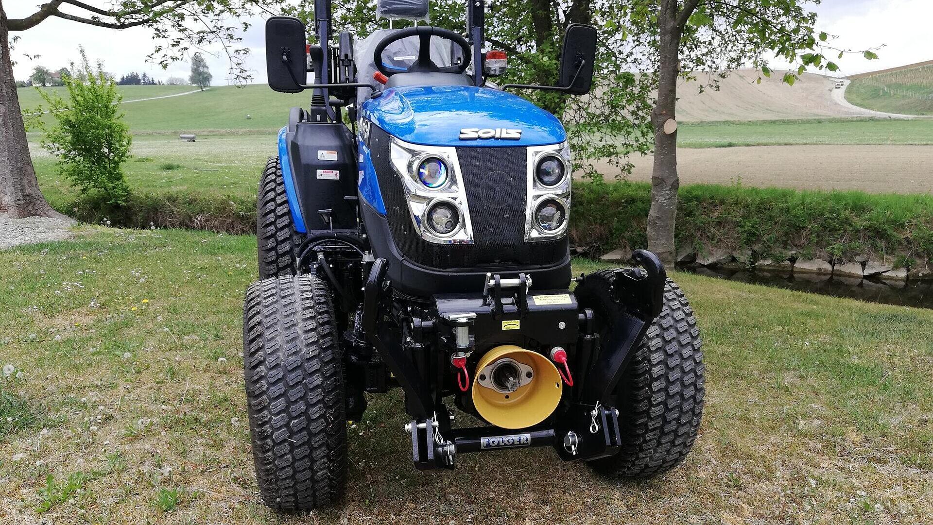 Ein blauer Solis-Traktor ist auf einer Wiese in der Nähe eines kleinen Baches und von Bäumen geparkt, während im Hintergrund Felder und ein gewundener Weg zu sehen sind. Das Foto zeigt die Vorderansicht des Traktors.