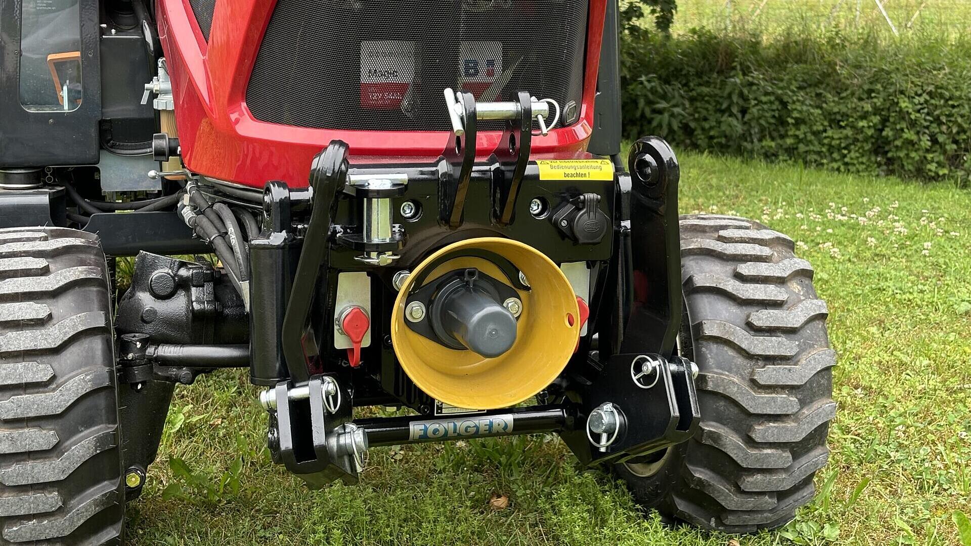 Nahaufnahme des vorderen Anbaugerätebereichs eines roten Traktors mit Zapfwelle und Hydraulikanschlüssen, mit großen Reifen und Gras und Ackerland im Hintergrund.