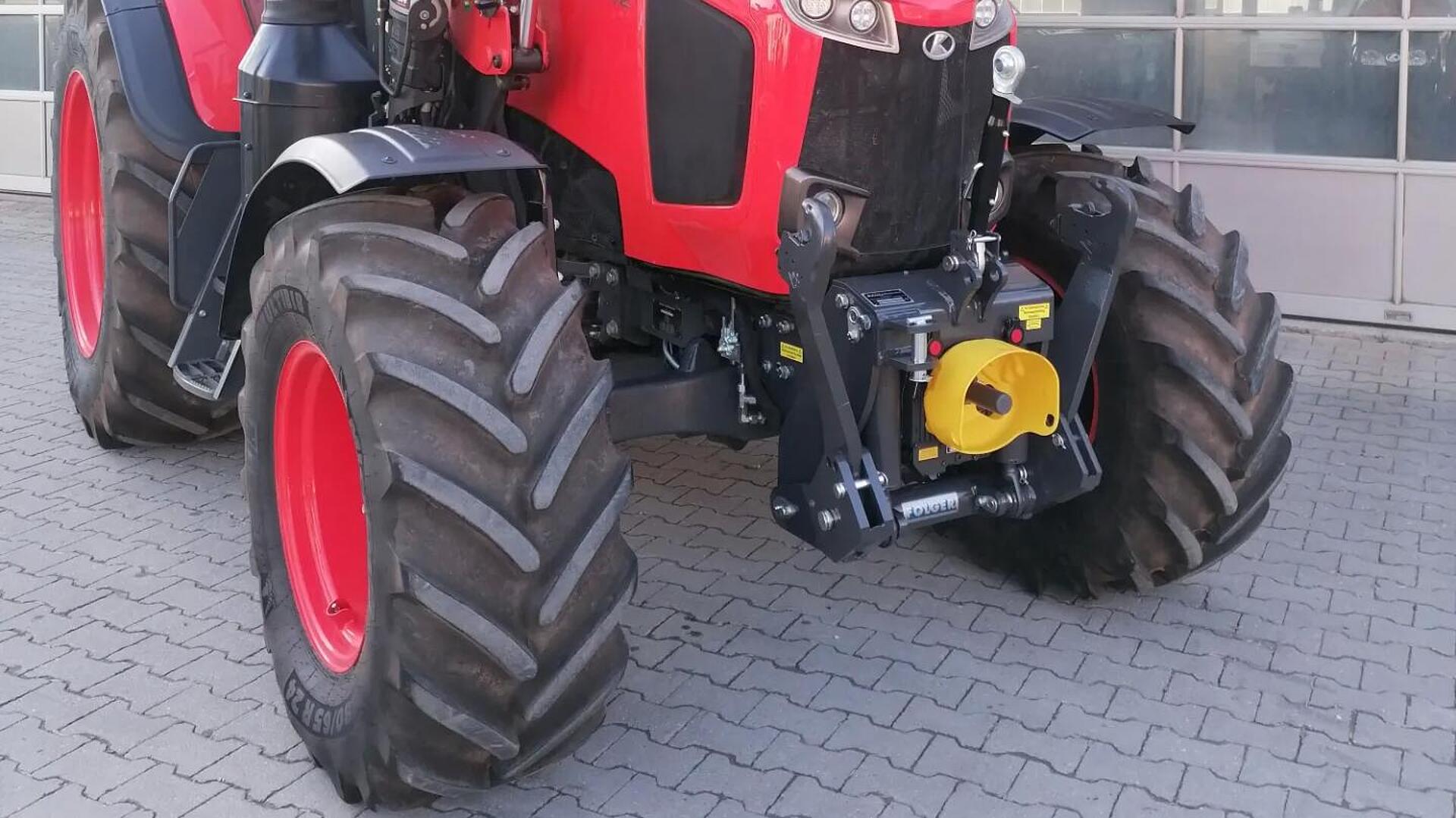 Ein roter Traktor mit großen schwarzen Reifen ist auf einer gepflasterten Fläche vor einem Gebäude mit großen Glastüren geparkt. Der Traktor ist nach vorne gerichtet und zeigt seine Scheinwerfer und den Frontanbau.
