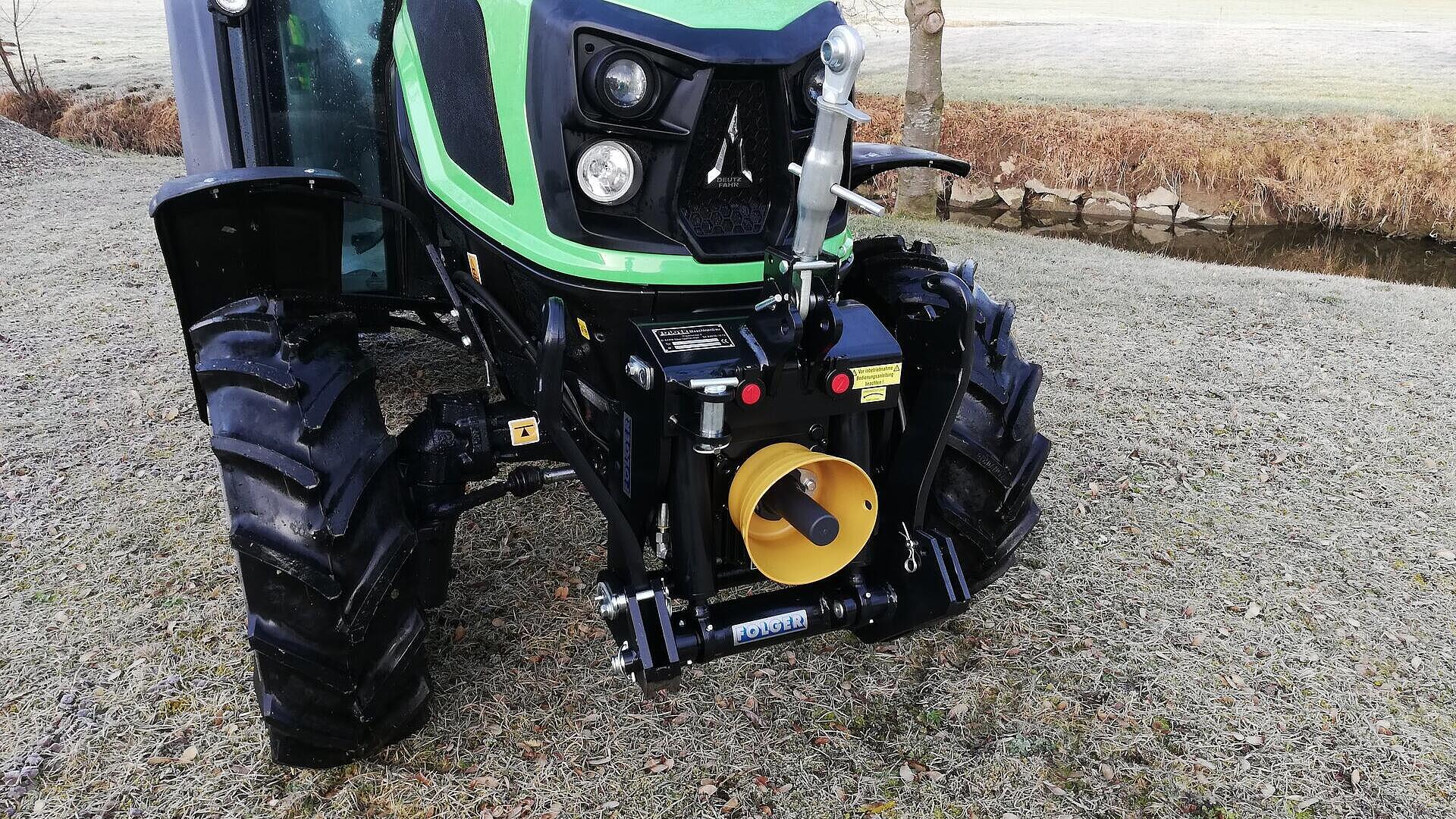 Ein grüner Traktor ist auf einer Wiese in der Nähe eines kleinen Baches geparkt, sein Frontanbau und die schwarzen Reifen sind deutlich zu sehen. Im Hintergrund sieht man einen kahlen Baum und weite Felder.