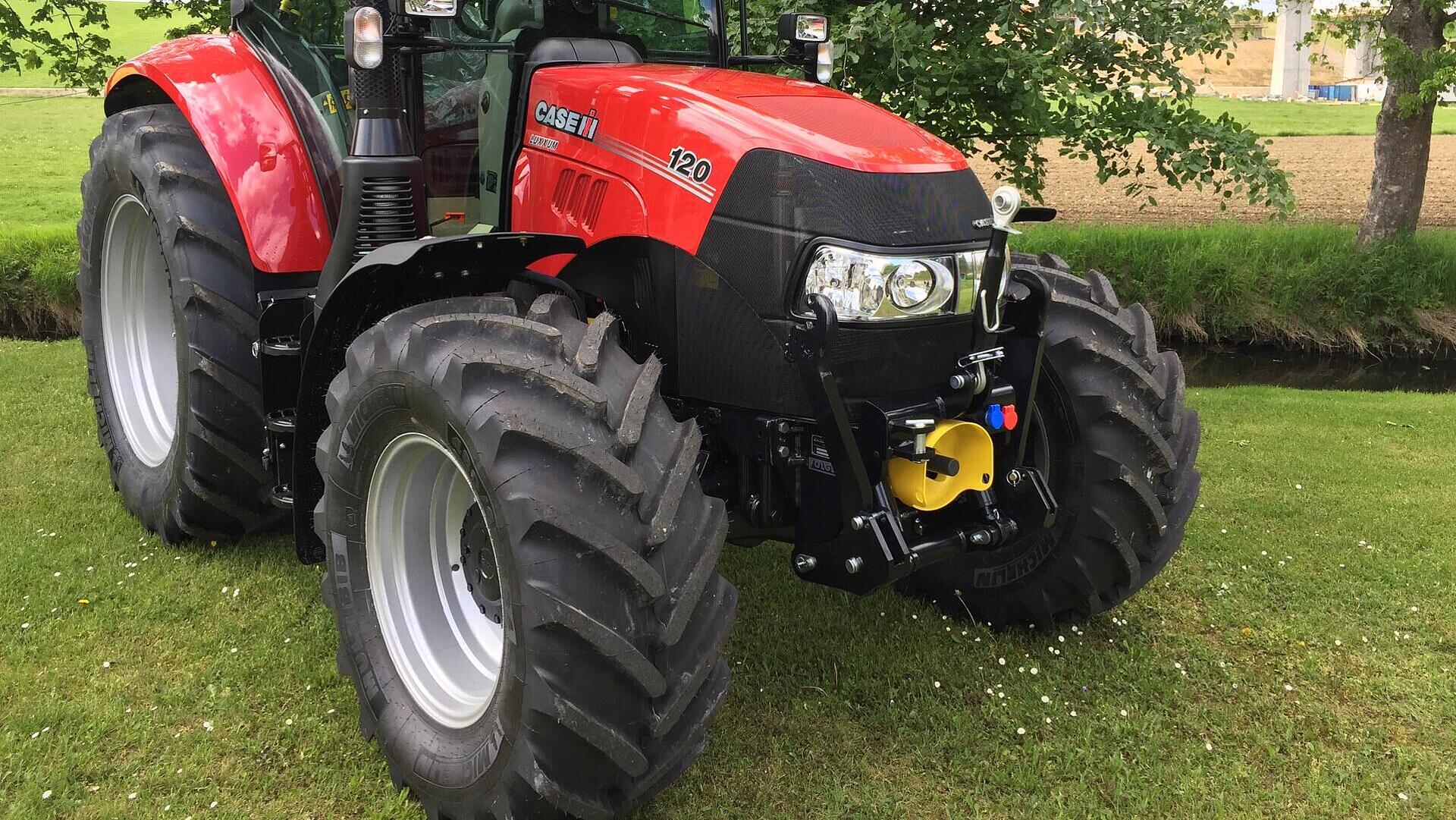 Ein roter Case IH 120 Traktor ist auf einer Wiese in der Nähe von Bäumen und Ackerland geparkt, mit großen Vorder- und Hinterreifen und sichtbaren Metalldetails unter einem teilweise bewölkten Himmel.